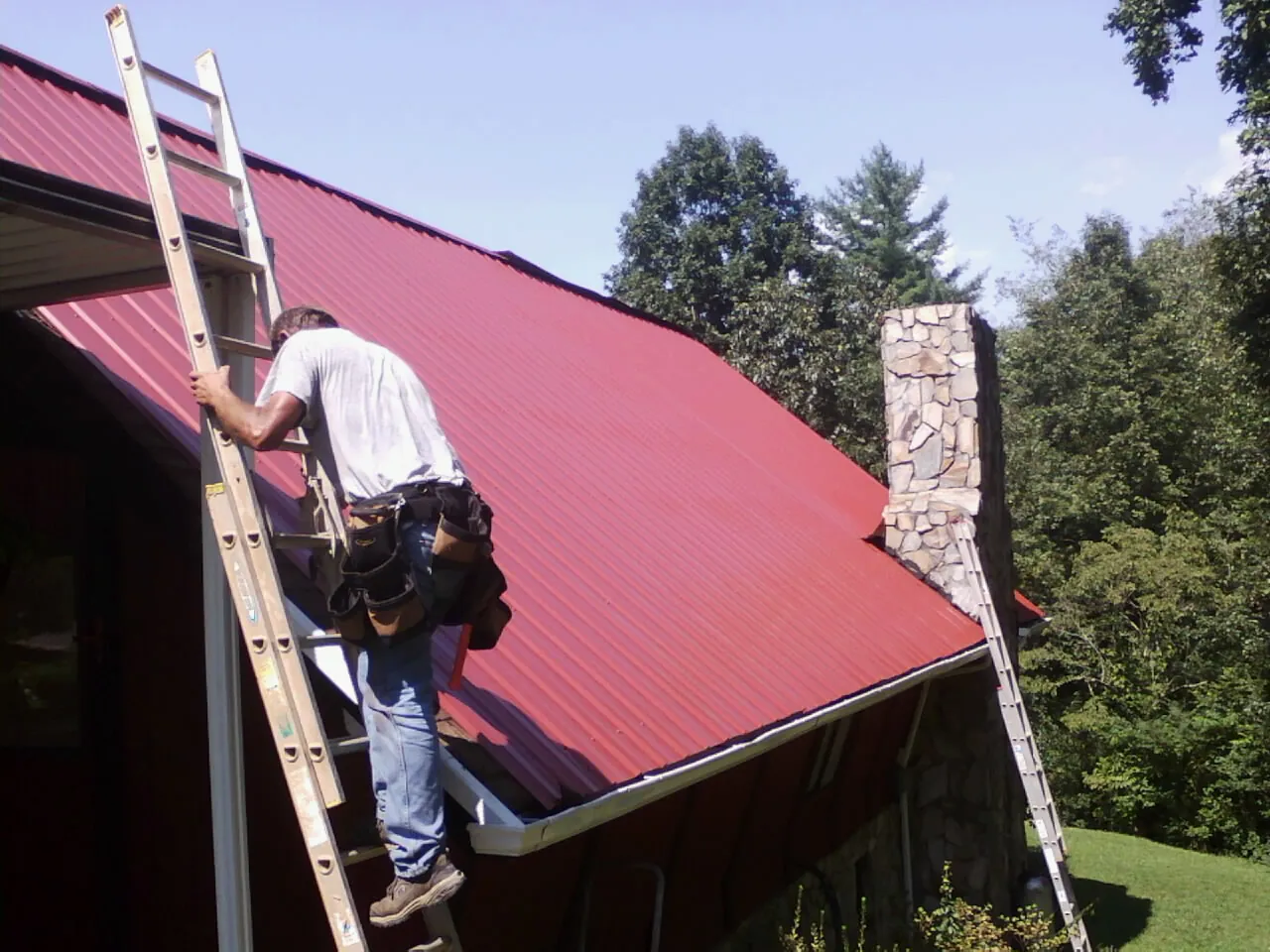 Roofer on a ladder beside a red metal roof and stone chimney — Soffit & Fascia Repair by Roofing Brothers in Fort Leavenworth